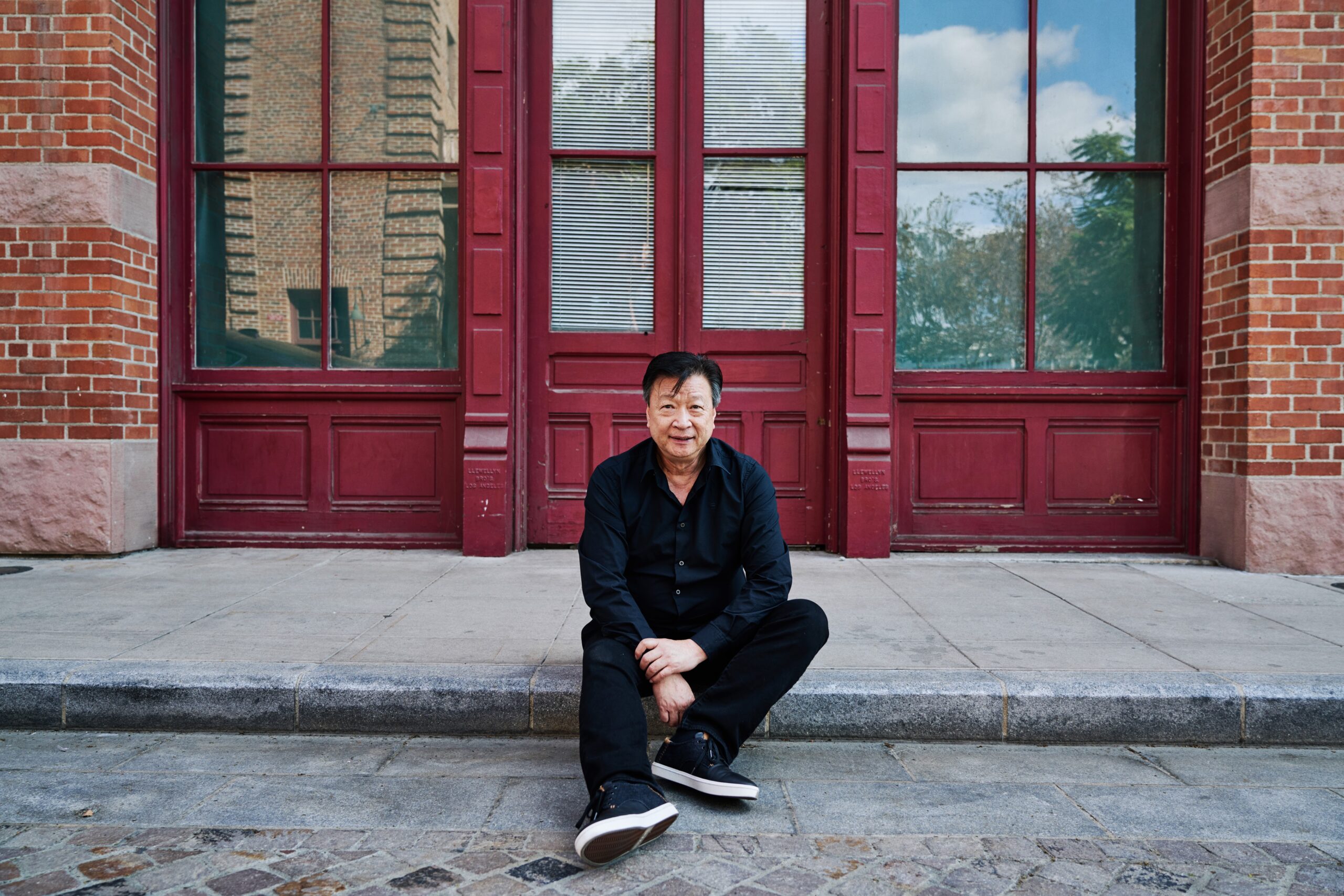 Tzi Ma seated on a curb in front of red doors