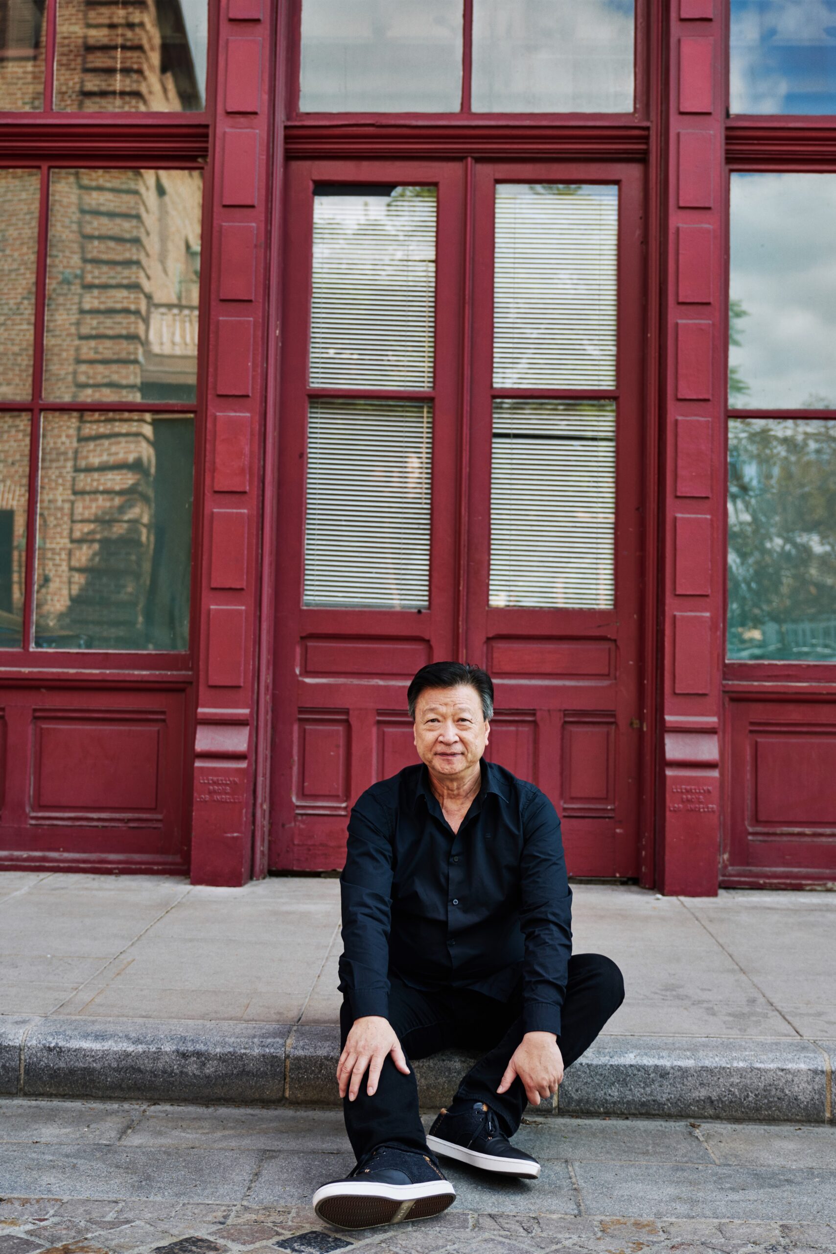 Portrait of Tzi Ma seated in front of tall red doors